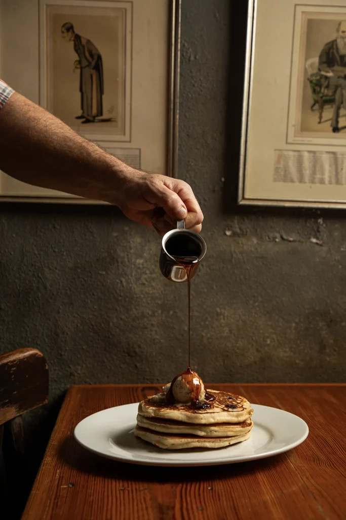 Hand pouring dark syrup onto pancakes with a scoop.