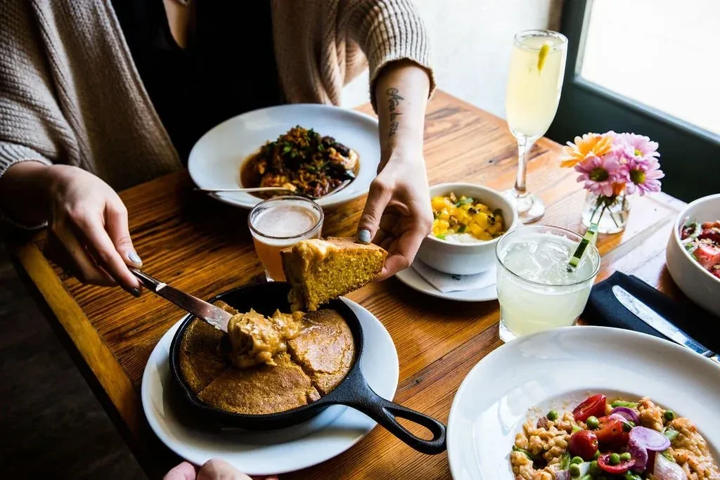 Hands lift cornbread from skillet on rustic table laden with food.