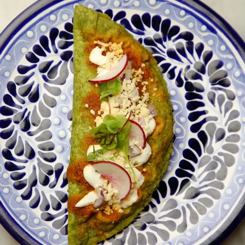 Green folded dish with sauce, cheeses, radish, herbs, on patterned plate.