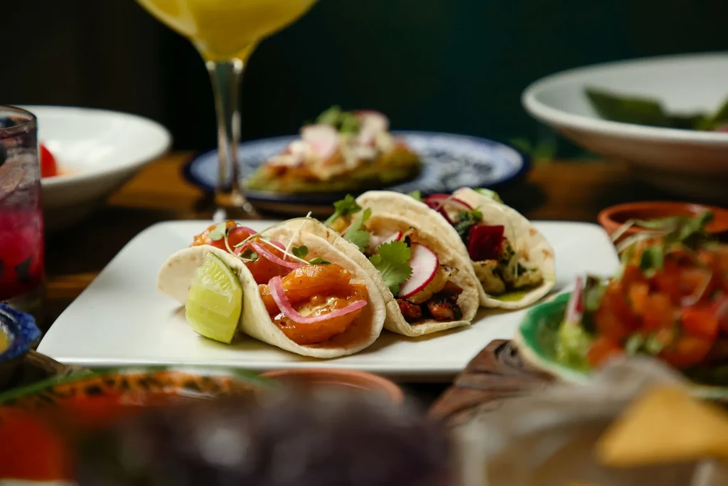 Close-up of three vibrant tacos with blurred drinks and food.