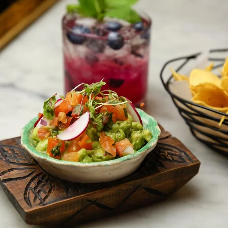 Guacamole bowl topped with tomatoes, radish, microgreens, near chips and drink.