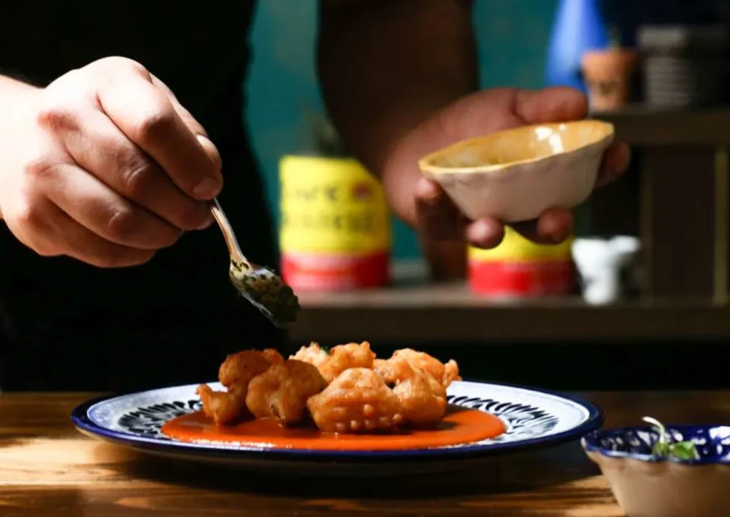 Hands drizzle green sauce onto golden fried fritters in orange sauce.
