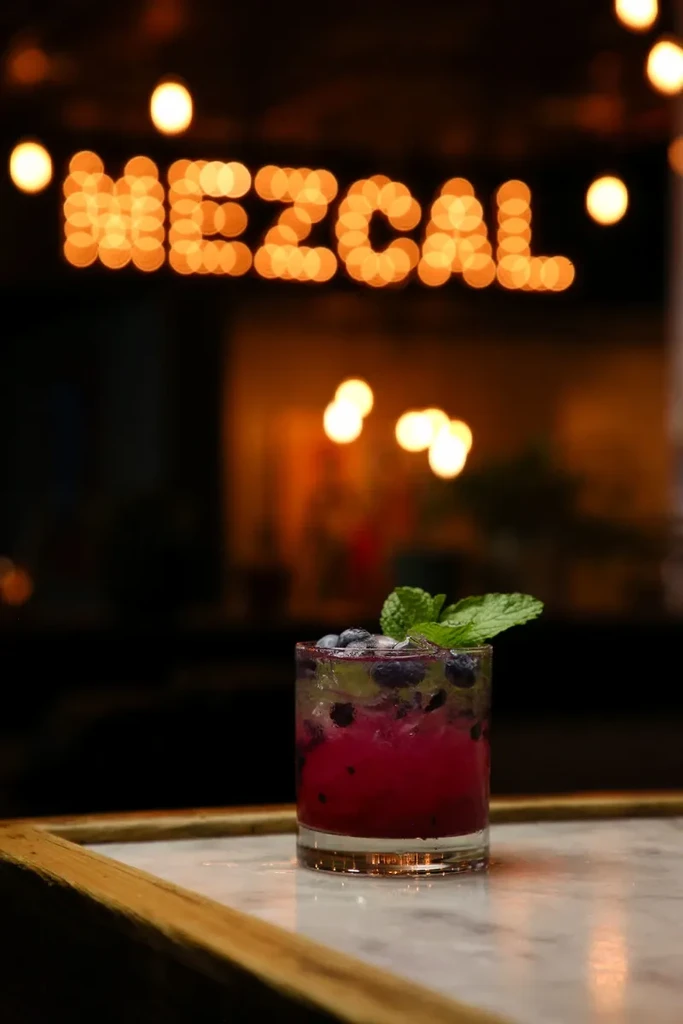 Magenta cocktail with blueberries, mint; "MEZCAL" sign in blurred background.