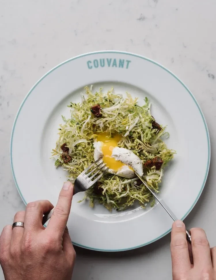 Hands cut a runny poached egg on a frisée salad with bacon.