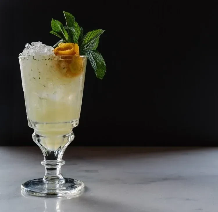 Light yellow cocktail in stemmed glass with crushed ice, mint, orange peel.