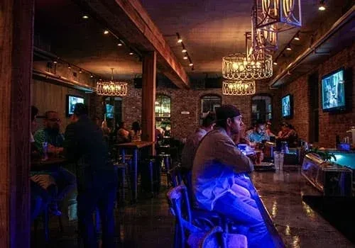 Warm, dimly lit bar interior with brick, wood, blue lights, and patrons.