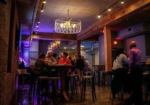 Lively industrial-rustic bar interior with people and warm, purple lighting.