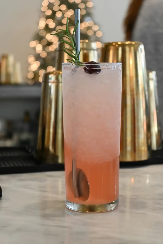 Layered pink and coral cocktail with rosemary, cherry, and blurred lights.