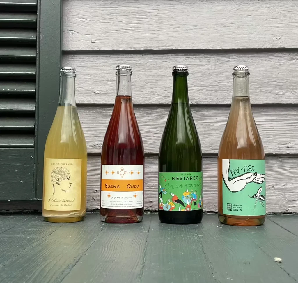 Four Pet-Nat wine bottles with colorful artistic labels on wood.