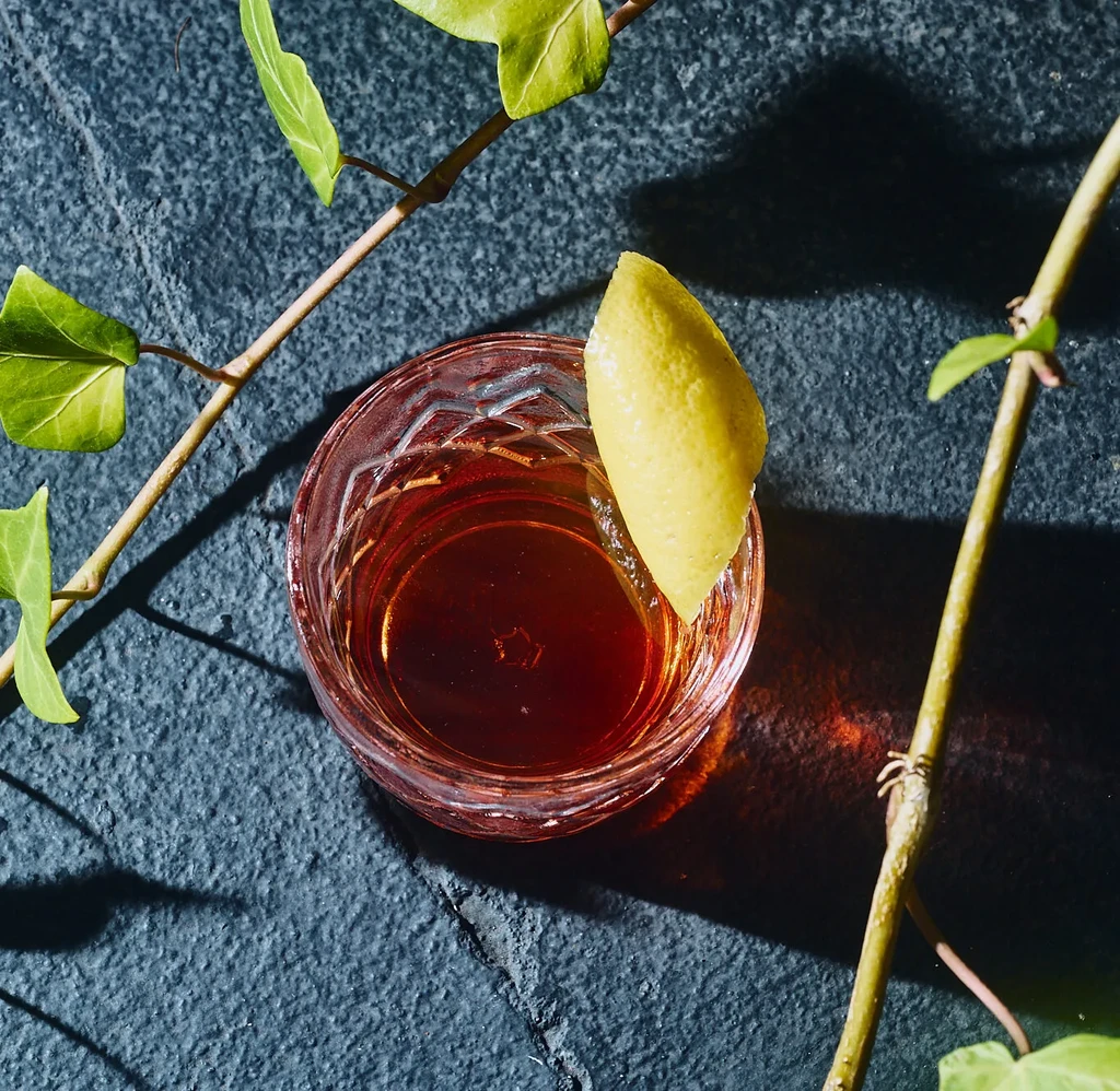 Top-down amber cocktail, lemon peel, textured glass on stone, with ivy.
