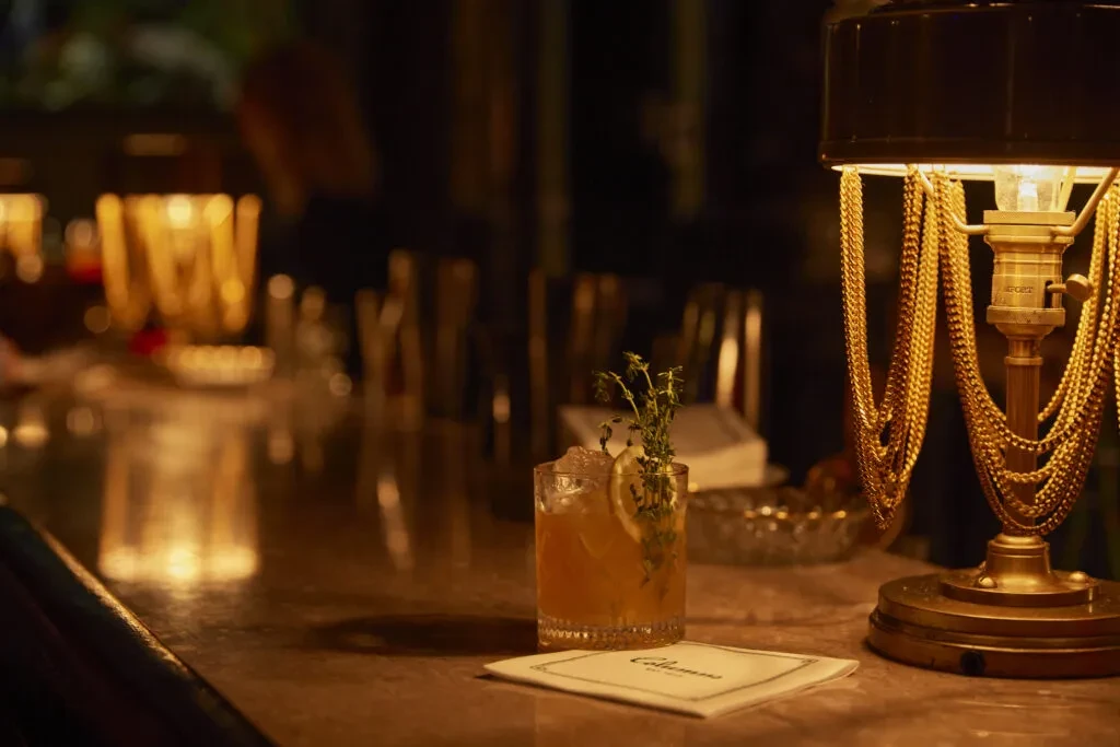 Lemon-garnished cocktail, ornate brass lamp, on a dim bar counter.