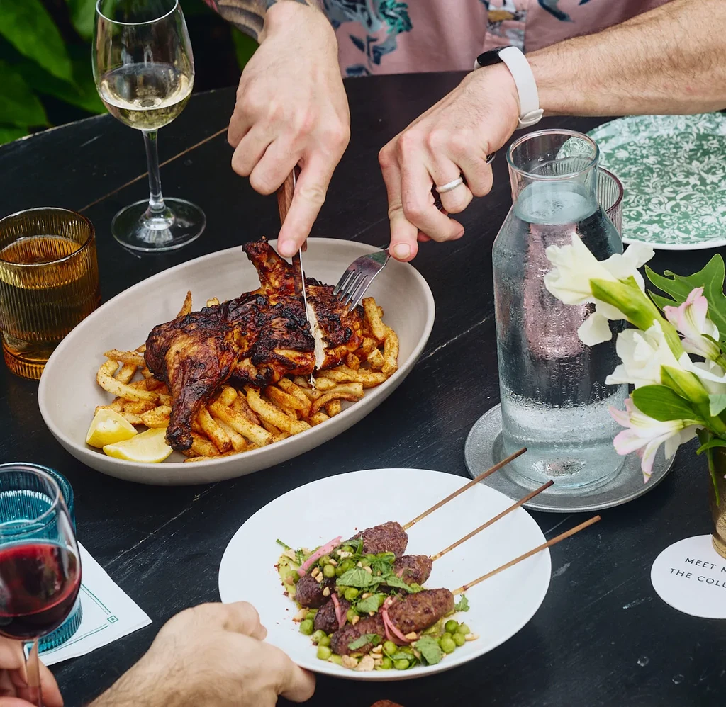 Hands carving chicken, kofta, fries, salad, and drinks on a restaurant table.