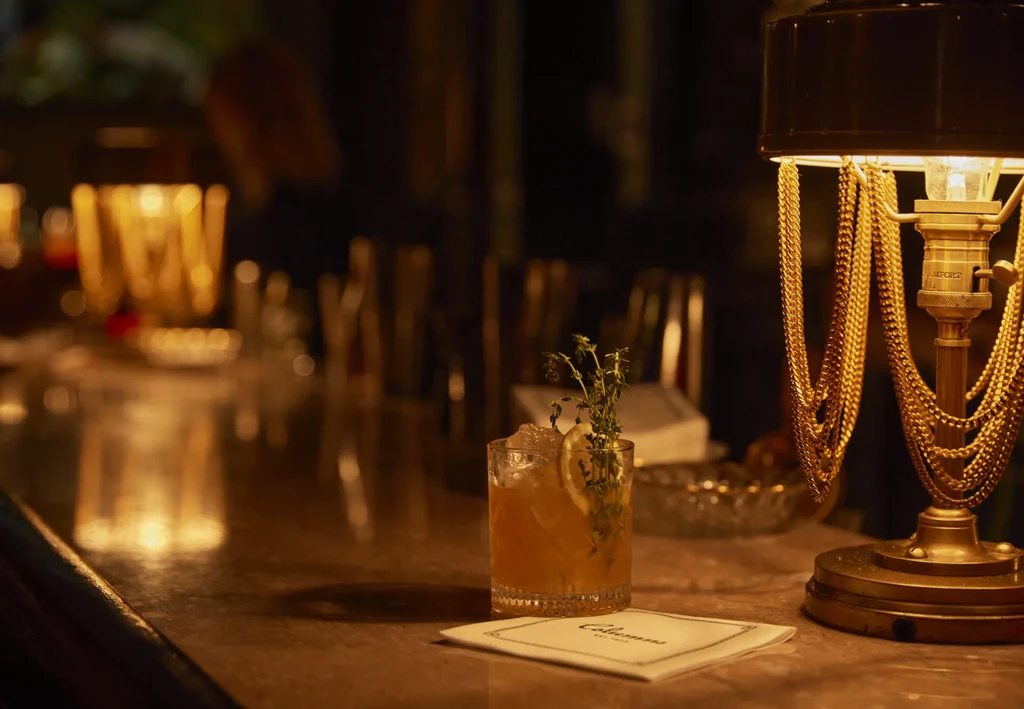 Amber cocktail with lemon and herbs on a dimly lit bar.
