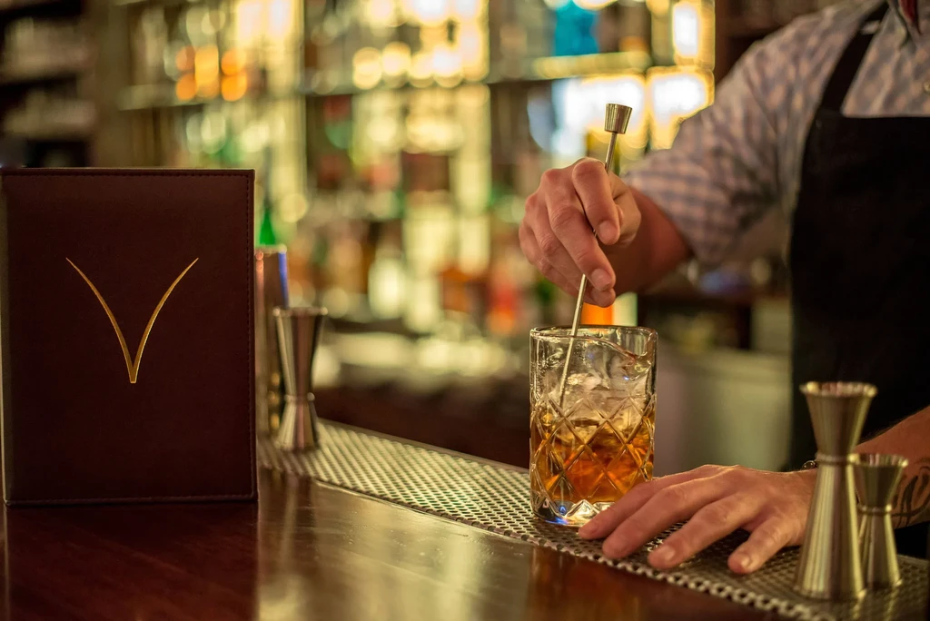 Bartender's hands stir amber cocktail in faceted glass with ice.