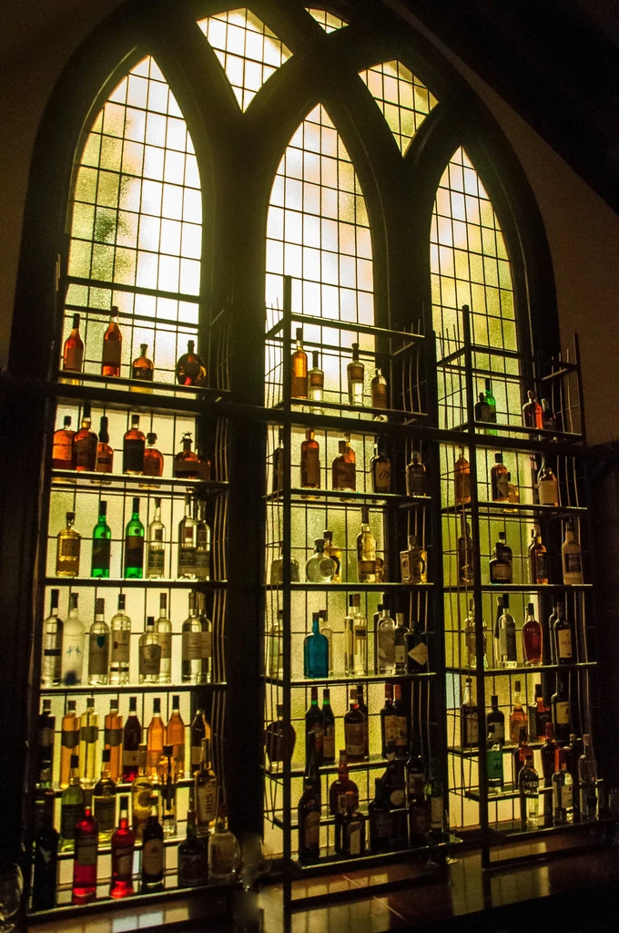 Liquor bottles on shelves in front of backlit, arched stained glass windows.