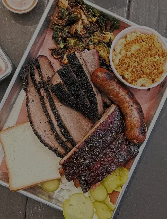 Overhead barbecue platter with brisket, ribs, sausage, mac and cheese, sprouts.