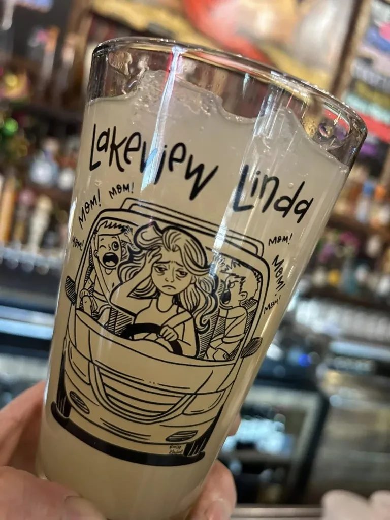 Hand holds a foamy drink in a glass with "Lakeview Linda" cartoon.