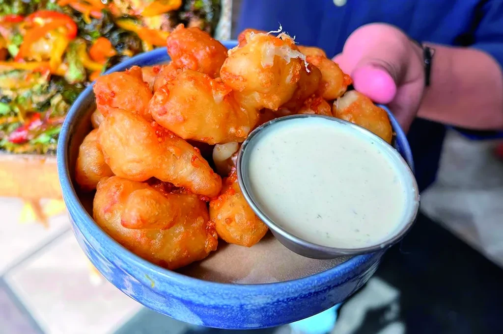 Person holds blue bowl of golden-fried cheese curds with dipping sauce.