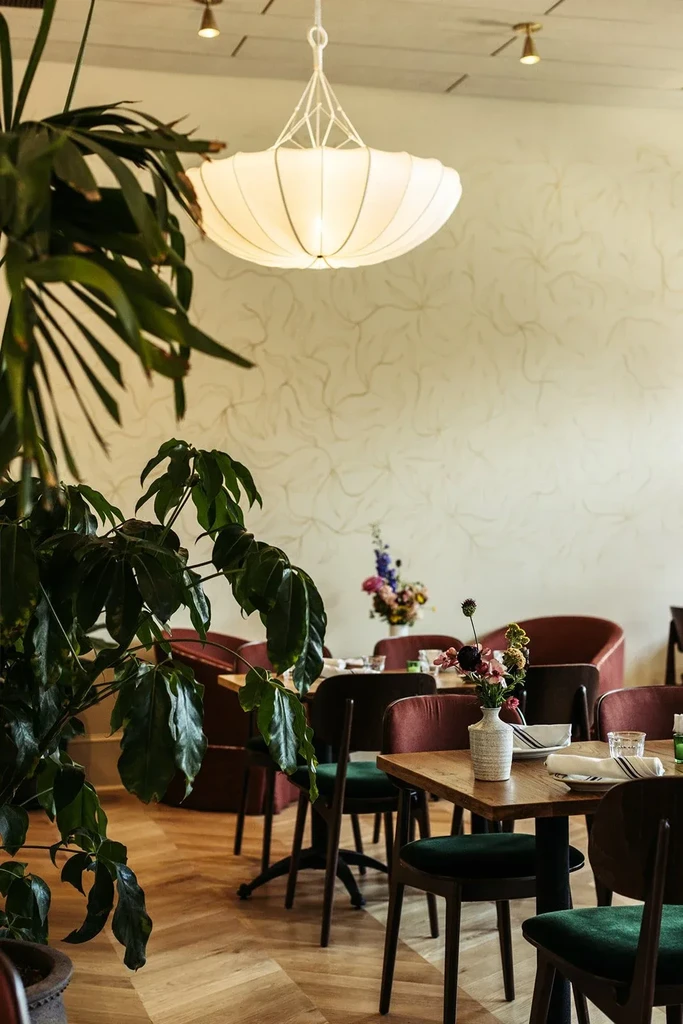 Bright restaurant interior with plants, wooden tables, and a large chandelier.