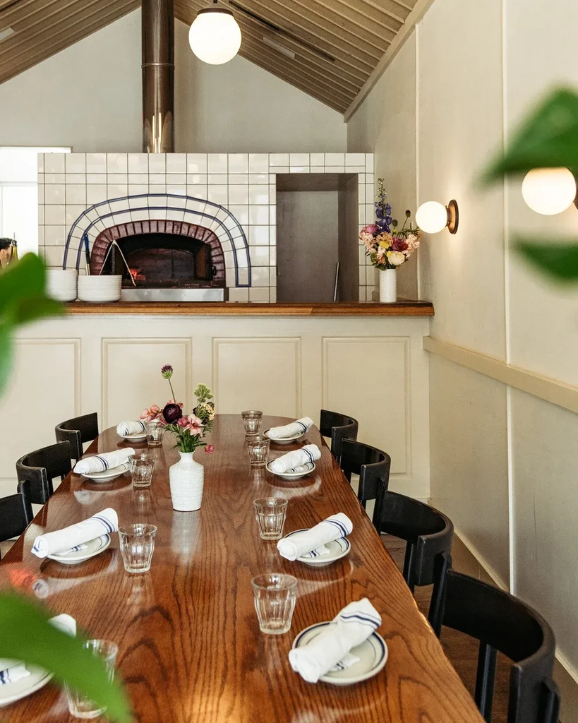 Rustic modern restaurant interior with long dining table and pizza oven.