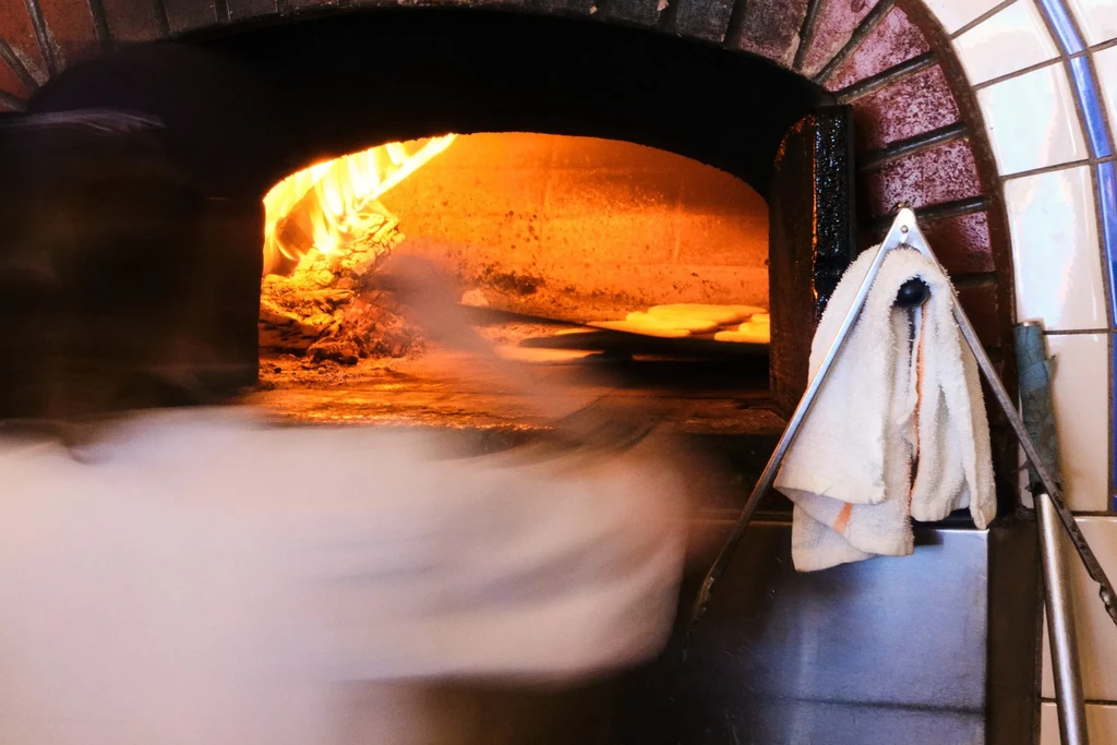 Glowing wood-fired oven baking pizzas, with flames and billowing smoke.