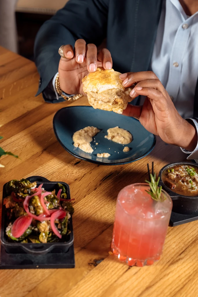 Person holds a creamy biscuit sandwich, roasted Brussels sprouts, and a cocktail.