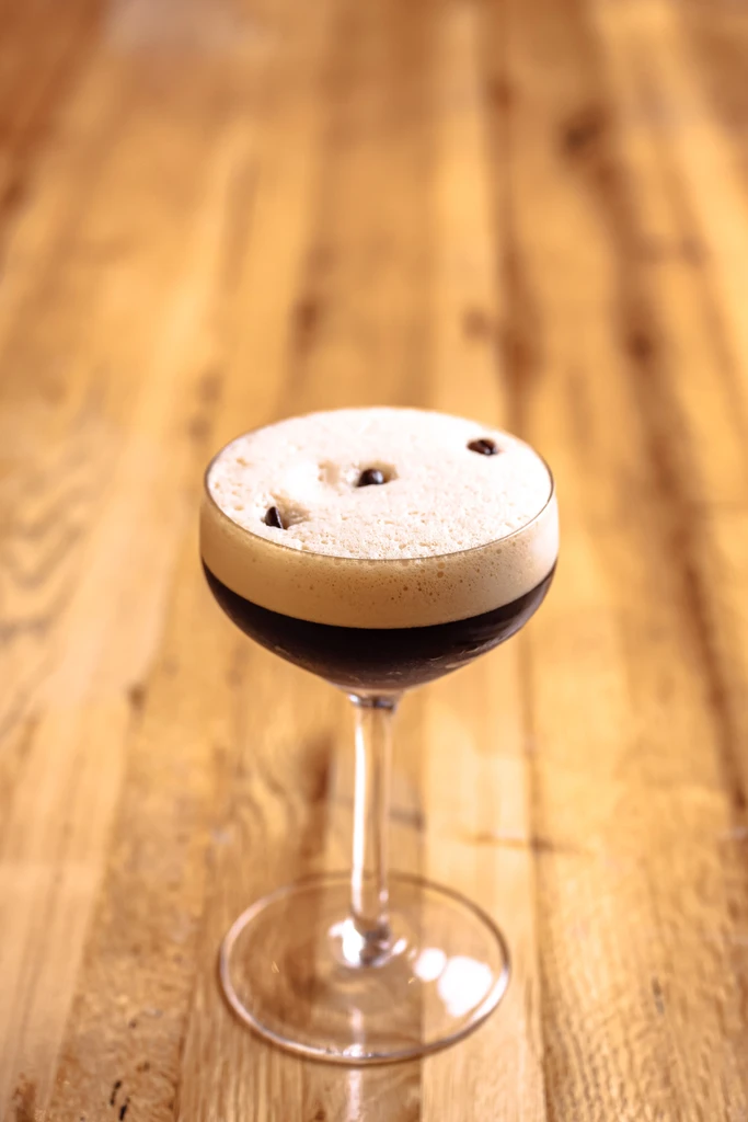 Dark cocktail with frothy foam and coffee beans in a coupe glass.