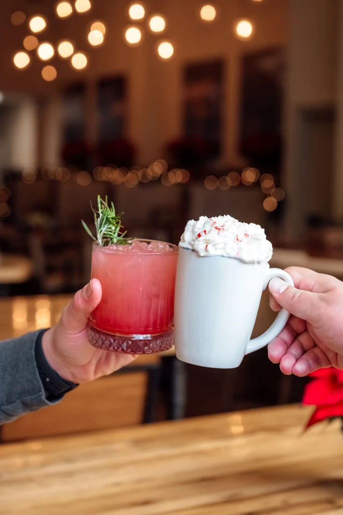 Hands toast a pink cocktail with rosemary and a candy cane mug.