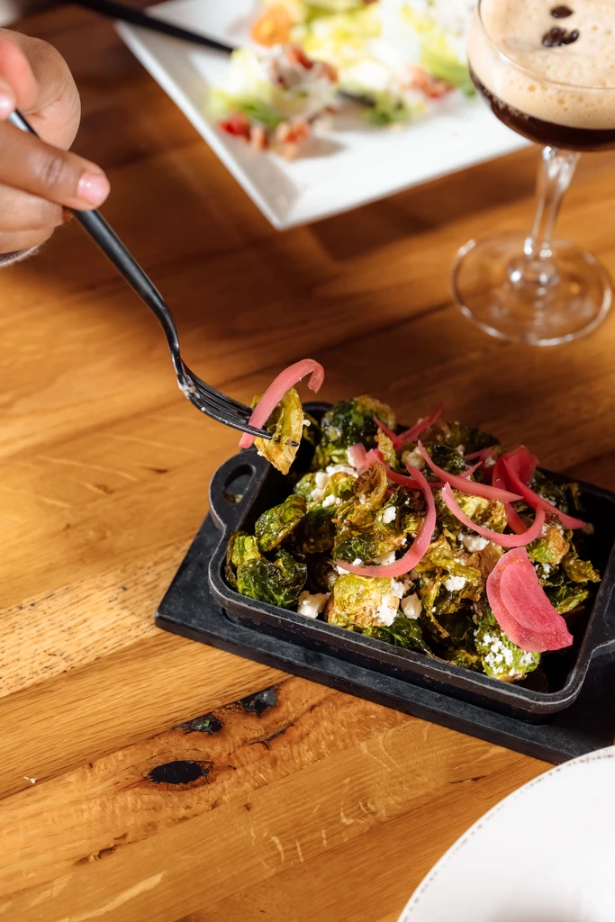 Hand forks roasted Brussels sprouts from cast iron dish with drink.