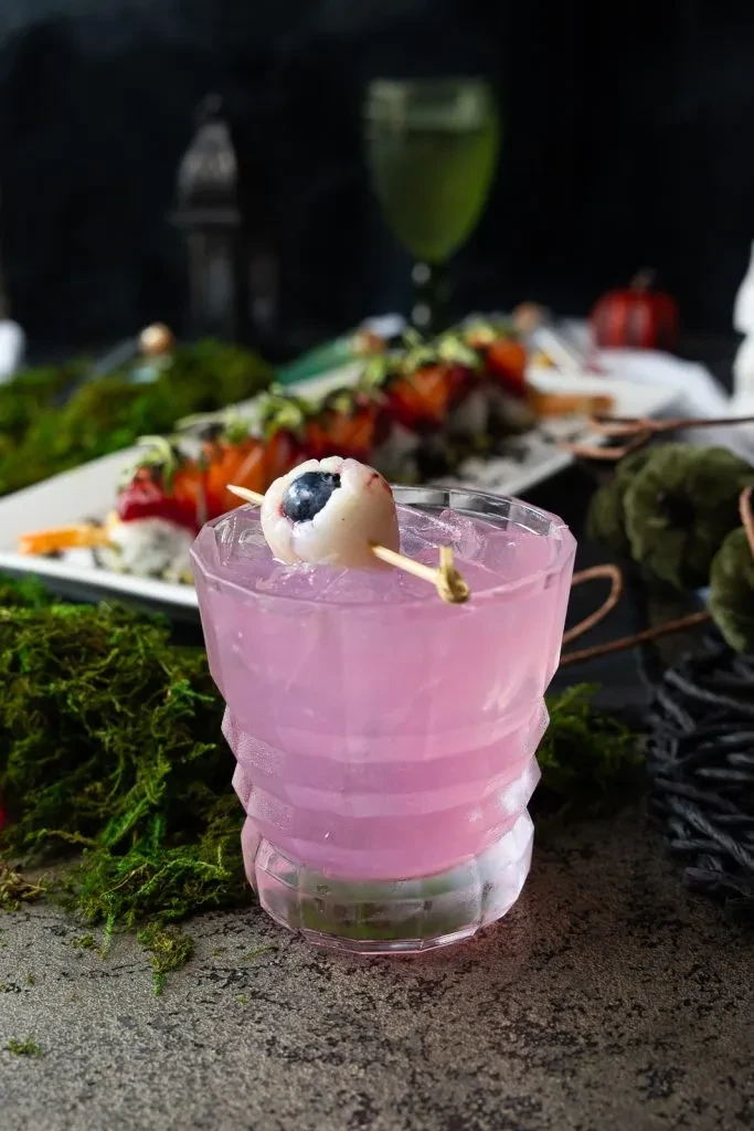 Pink cocktail in faceted glass with lychee eyeball, ice, Halloween theme.
