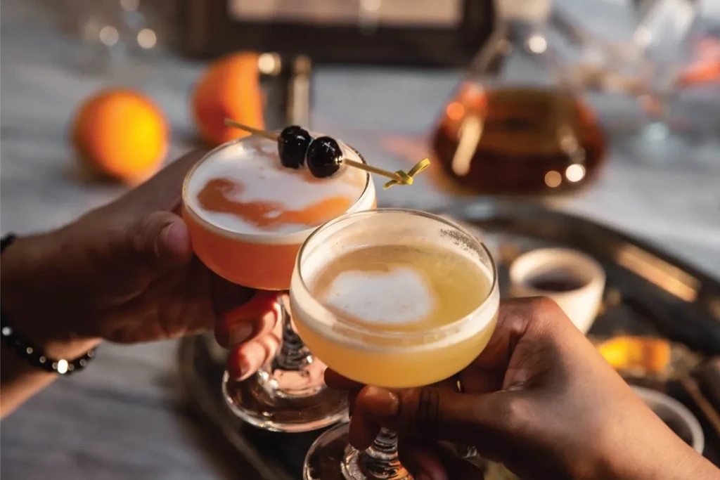 Two hands clink two frothy cocktails: one red with cherries, one yellow.
