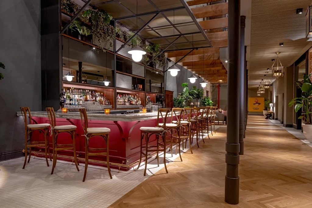 Modern bar with red counter, liquor shelves, hanging plants, and long hallway.