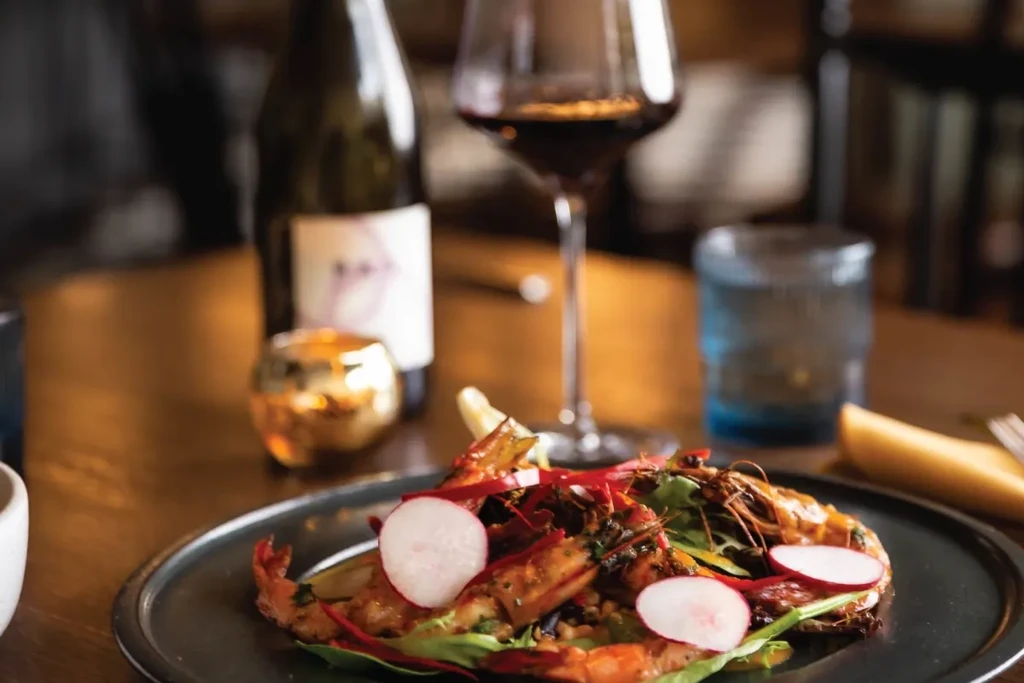 Dark plate with grilled prawns, greens, red strips, and white radish.