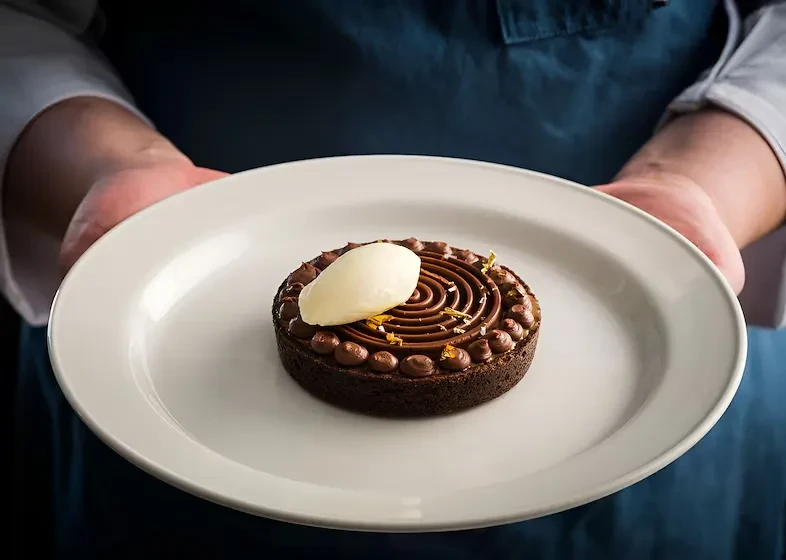 Hands holding a dark chocolate tart with ice cream and gold flakes.