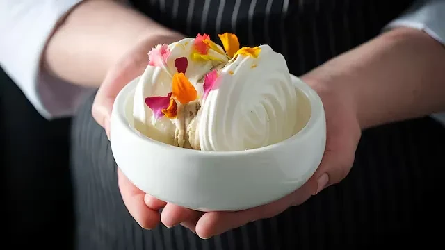 Chef presents swirled white dessert with edible flowers in a bowl.