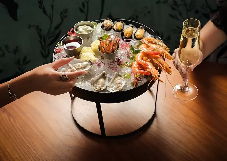 Hands with champagne, reaching for oysters on a tiered seafood platter.