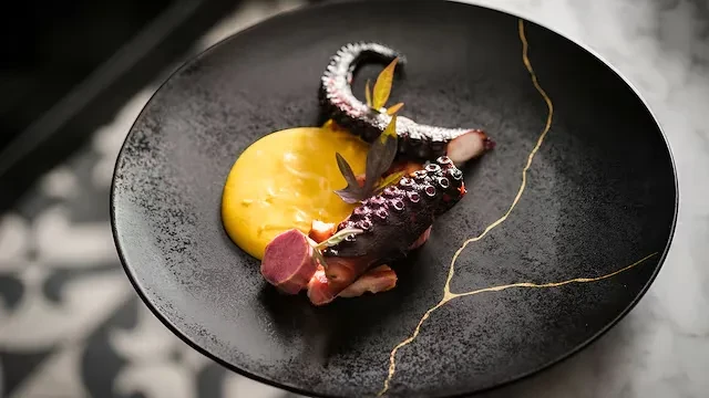 Grilled octopus with yellow puree, root vegetables, and dark leaves.