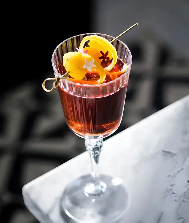 Dark cocktail in fluted glass, garnished with citrus twists and flowers.