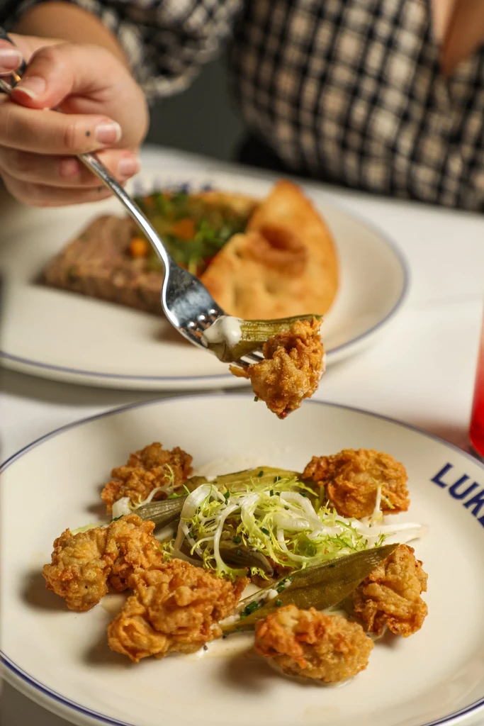 A hand lifts fried golden food and okra with sauce from a plate.