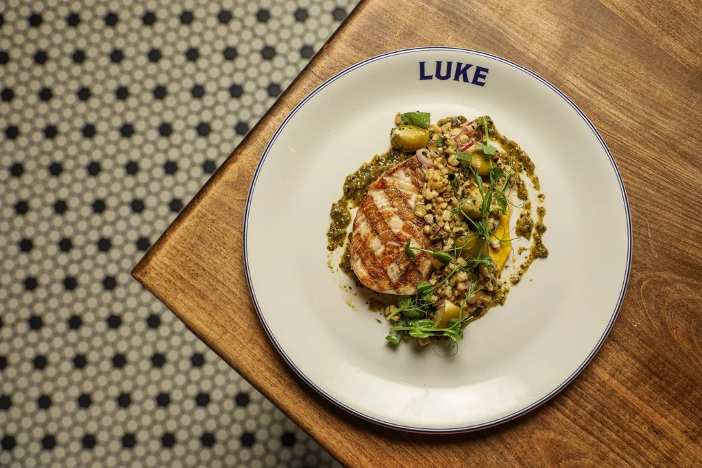 Grilled protein with green sauce, grains, olives, on 'LUKE' branded plate.
