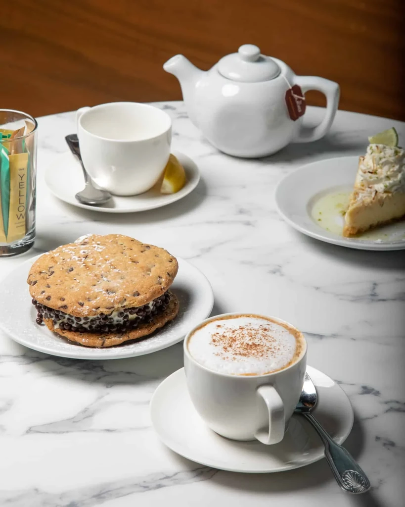 Marble tabletop with cappuccino, cookie sandwich, cheesecake slice, and tea.
