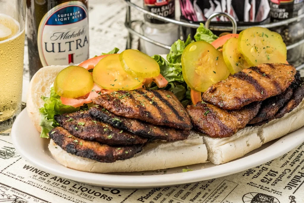 Overflowing grilled meat sandwich with lettuce, tomatoes, pickles, and beer.