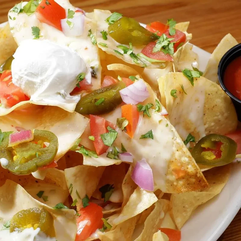 Plate of nachos with melted cheese, vegetables, sour cream, and salsa cup.