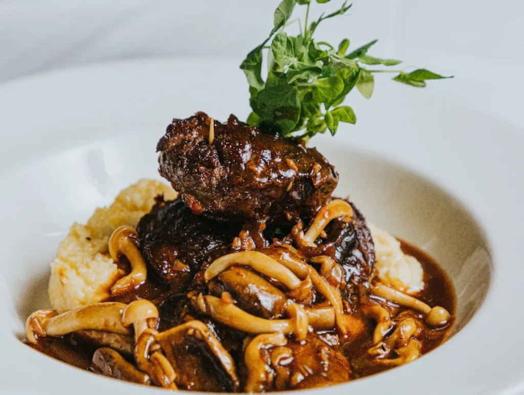Glistening braised meat, mushrooms, herbs, and creamy polenta in white bowl.