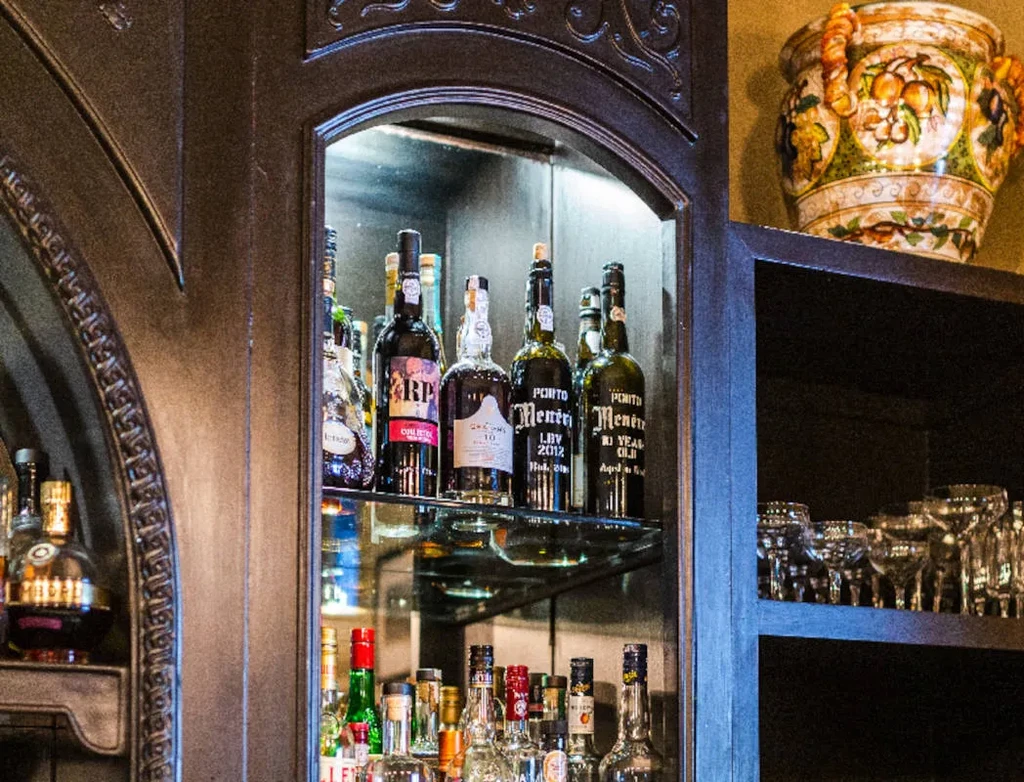 Dark wooden bar cabinet featuring bottled spirits, glassware, and ornate vase.