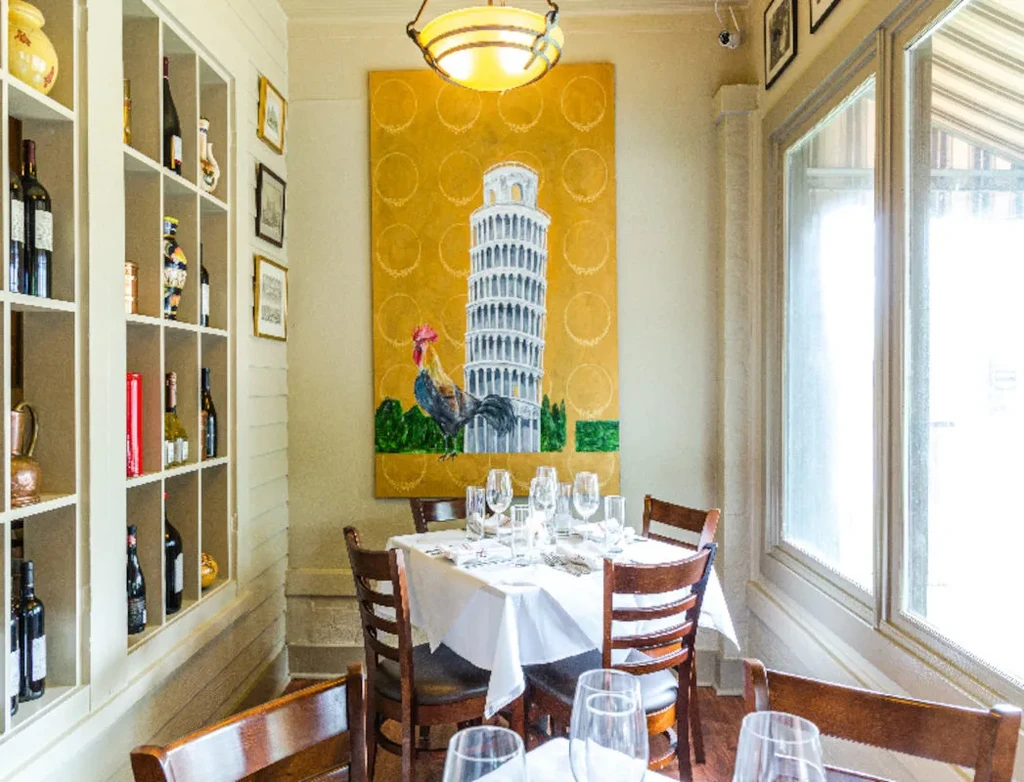 Italian restaurant interior with set table, wine display, and Pisa painting.