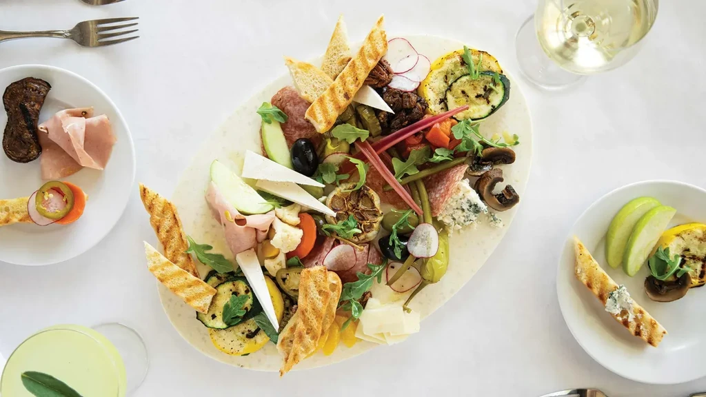 Overhead, overflowing charcuterie board with diverse foods, plates, and drinks.
