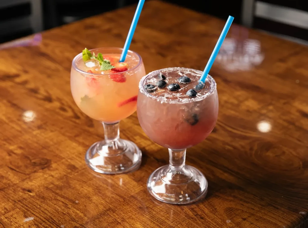 Two vibrant cocktails with fruit garnishes and straws on a wooden table.