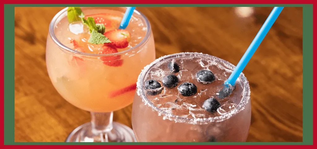 Two colorful cocktails in stemmed glasses with fruit, ice, and straws.