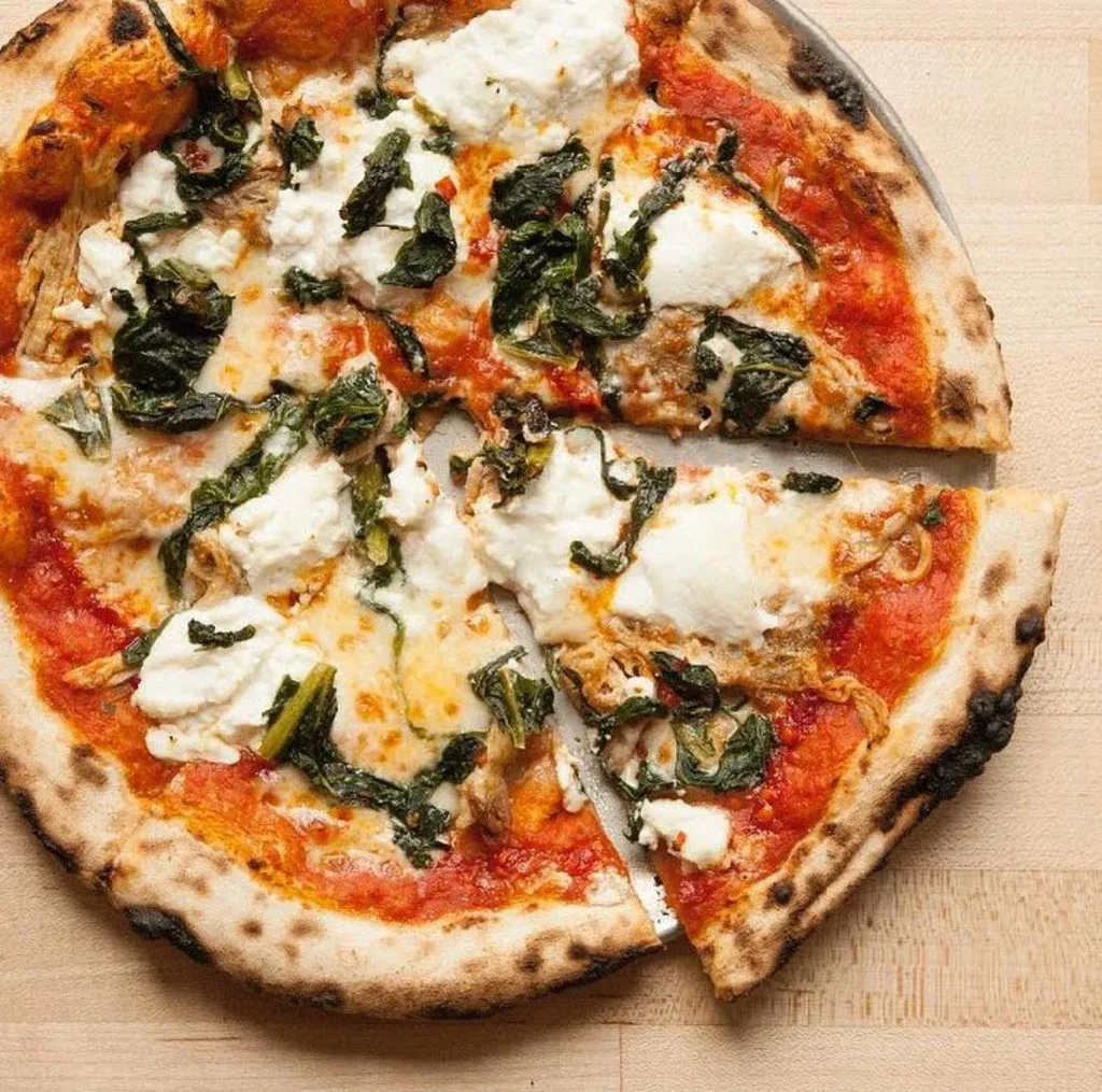 Wood-fired pizza with tomato, greens, melted cheese, and a cut slice.